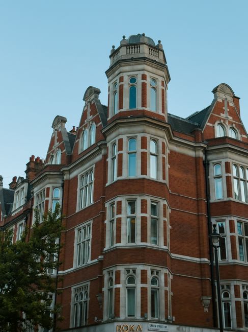 A tall, historic red brick building with multiple stories and ornate architectural details, including rounded bay windows framed with white trim, decorative stone accents, and a turreted top with a small balcony, located on a street corner under a clear blue sky. In the foreground, a black street lamp and a partially visible leafy tree are present. The building's ground floor features a sign reading 'ROKA' and the address 'Green Street, SW3,' indicating it is situated in Chelsea. This setting suggests a preserved colonial or Victorian-style residence, typical of a high-end residential area, often associated with house removals and home relocation services such as those offered by Man with Van Chelsea. The image captures the exterior facade during daylight, emphasizing its historic charm and prominent position in an urban environment, relevant to furniture transport and moving logistics.