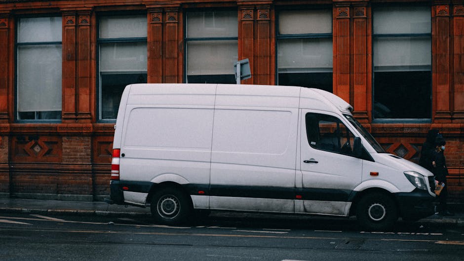 A white cargo van parked parallel to a red brick building with ornate architectural details, situated on a city street. The van's side door is visible, with no visible markings or signage. Two pedestr