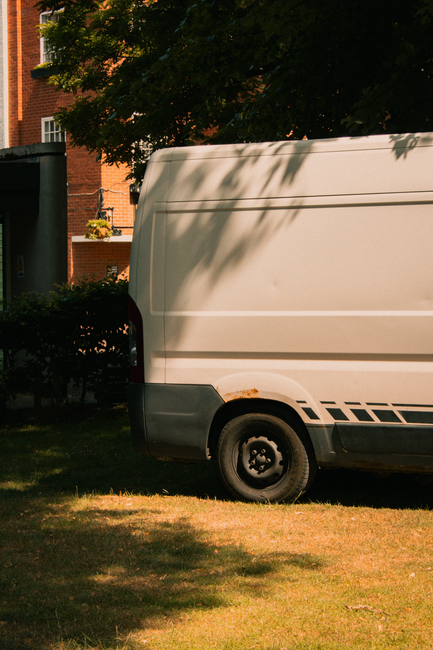 A white cargo van parked parallel to a red brick building with ornate architectural details, situated on a city street. The van's side door is visible, with no visible markings or signage. Two pedestr