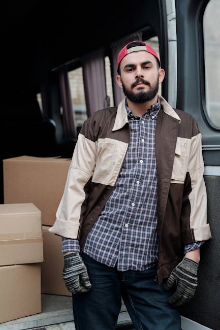 A young man with a beard, wearing a black cap and a white polo shirt, stands inside the open rear doors of a cargo van loaded with multiple cardboard boxes of various sizes, some sealed with packing t