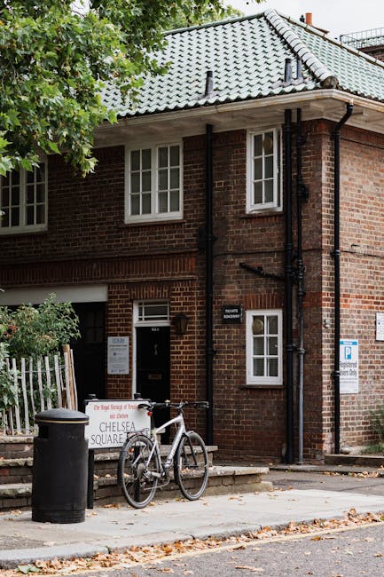 The exterior of a two-storey brick residential building in Chelsea, with a grey tiled pitched roof and white-framed windows. A black metal mailbox is mounted beside the dark doorway, which is partially open, indicating ongoing house removals. In front of the building, a silver bicycle with a black saddle and handlebars is parked against a waist-high white metal fence. A black trash bin is positioned on the pavement nearby. The scene includes a temporary sign for Chelsea Square, related to the relocation service provided by Man with Van Chelsea, illustrating the context of home relocation or furniture transport. The pavement is scattered with fallen leaves, and the area is partially shaded by overhanging green tree branches, with natural daylight illuminating the scene. This setting captures a typical residential street environment in the process of moving or packing activities.