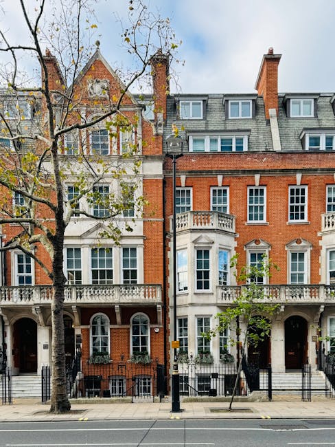 The image depicts a row of three-storey residential buildings in Chelsea, featuring red and white brick facades with decorative architectural elements such as arched windows, balustrades, and ornate window surrounds. The buildings are situated directly on the pavement, with a leafless tree partially obscuring the view on the left. In front of the buildings, there is a black metal fence and a paved sidewalk. A black post and a tree planter are visible on the pavement, indicating a typical urban street scene. The sky is partly cloudy with patches of blue visible. This setting provides a suitable backdrop for house removals or furniture transport, aligning with the services offered by Man with Van Chelsea, specializing in home relocation and moving logistics near Sloane Square, CHELSEA.